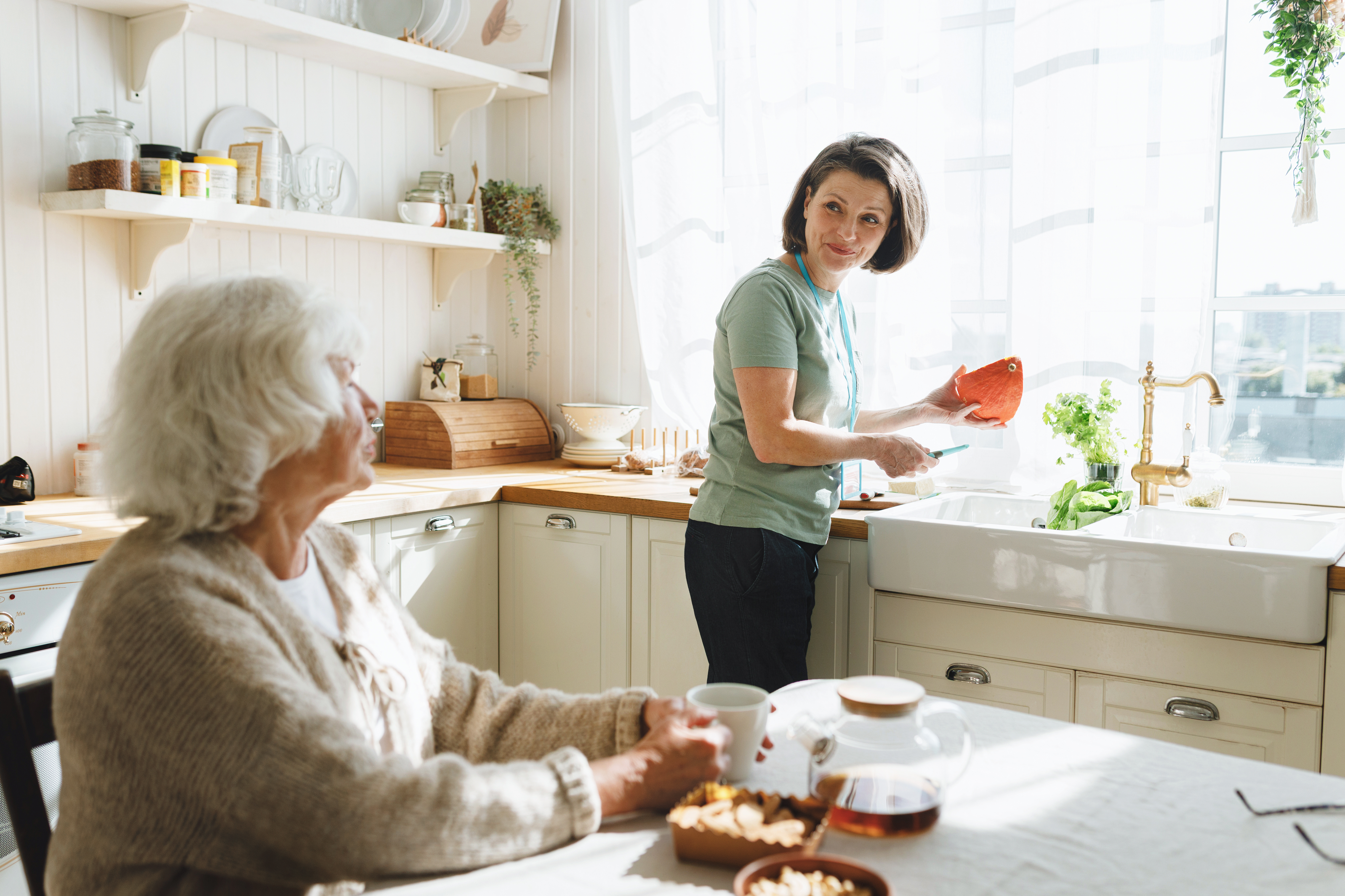 A caregiver and an older woman share a quiet, unhurried moment together in a home kitchen.